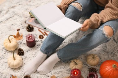 Arka plan olarak beyaz ekose üzerinde kitap, mum ve balkabağı ile sıcak çoraplar içinde kadın bacaklar