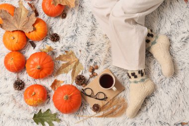 Bir fincan çay, sonbahar yaprakları ve arka plan olarak beyaz ekoseli balkabaklarıyla sıcak çoraplar içinde kadın bacakları.