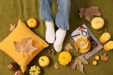 Sonbahar yaprakları, balkabakları ve arka plan olarak yeşil ekoseli bir fincan çayla sıcak çoraplar içindeki kadın bacakları.