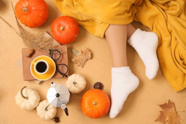 Sonbahar yaprakları, balkabakları ve bej arka planda bir fincan kahveyle sıcak çoraplar içindeki kadın bacakları.