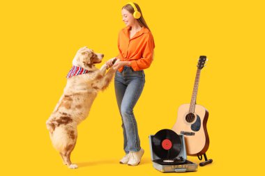 Kulaklıklı genç bir kadın sarı arka planda Avustralyalı çoban köpeği ve pikapla dans ediyor.