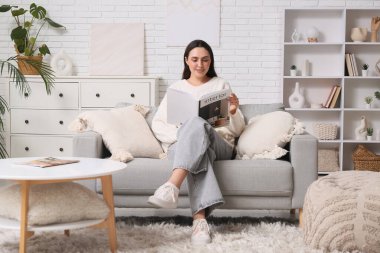 Evdeki kanepede iç tasarım dergisi okuyan genç bir kadın.