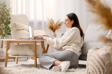 Evdeki masada dizüstü bilgisayar kullanan genç bir kadın.