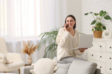 Evde dizüstü bilgisayarı olan genç bir kadın cep telefonuyla konuşuyor.