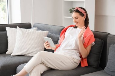 Tablet bilgisayarlı genç bir kadın evdeki kanepede sohbet ediyor.