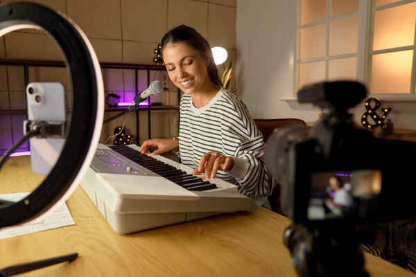 Female music blogger playing synthesizer while recording video at home in evening