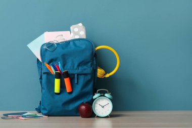 Mavi duvarın yanındaki ahşap masada farklı kırtasiye malzemesi ve alarm saati olan renkli okul çantası.