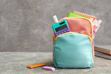 Duvarın yanındaki grunge masasında not defterleri, hesap makineleri ve işaretleyicilerle dolu renkli okul çantası.