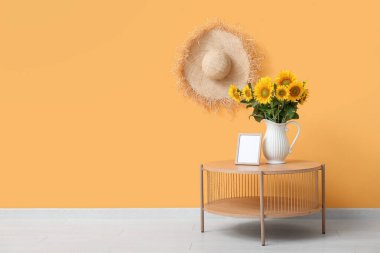 Güzel ayçiçekli vazo, boş çerçeve ve turuncu duvarda asılı hasır şapka.