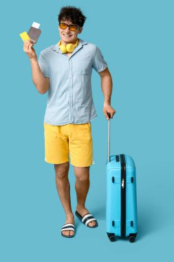Güneş gözlüklü genç adam mavi arka planda bavul, pasaport ve kredi kartıyla