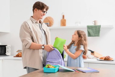 Babasıyla birlikte sırt çantasına defter koyup mutfaktaki okul çantasına koyan küçük bir kız.