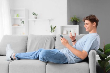 Evdeki kanepede bir bardak süt ve cep telefonuyla yatan genç adam.