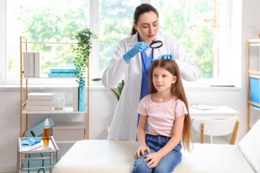 Büyüteci olan kadın doktor klinikte küçük kızın kafasında pediküloz olup olmadığını kontrol ediyor.