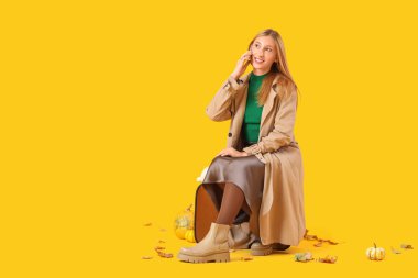 Cep telefonuyla konuşan genç ve güzel bir kadın balkabağı ve sonbahar yapraklarıyla sarı arka planda.