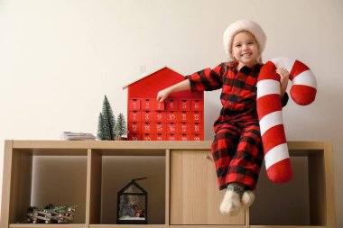 Pijamalı küçük bir kız ahşap rafta oturuyor. Noel takvimi ve ışık duvarına yazılmış mektuplar var.