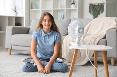 Teenage girl sitting on floor near electric fan in living room clipart