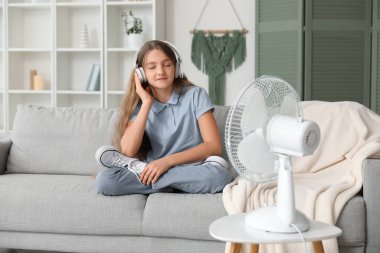 Teenage girl sitting on sofa near electric fan and listening music in living room clipart