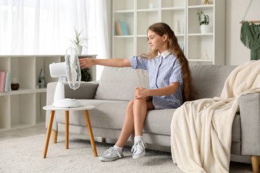 Oturma odasındaki elektrikli vantilatörün önünde oturan genç kız.