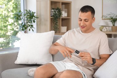 Nabız oksimetresi olan sportif genç adam evdeki kanepede oturuyor.