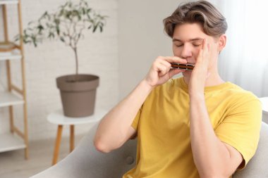 Genç adam evdeki koltukta mızıka çalıyor.