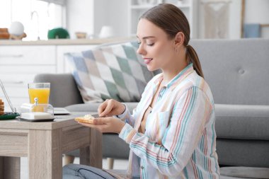 Güzel, genç ve mutlu bir kadın, elinde bir bardak meyve suyuyla, evde kızarmış ekmeğin üzerine taze tereyağı sürüyor.