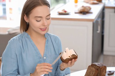 Güzel, genç ve mutlu bir kadın evde mutfakta tereyağlı lezzetli bir dilim ekmek yayıyor.