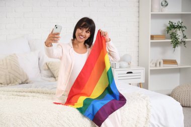 LGBT bayrağıyla güzel genç bir kadın selfie çekiyor ve yatak odasında oturuyor.