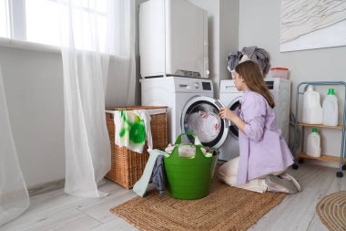 Çamaşır sepetli genç bir kadın çamaşır makinesine çamaşır koyuyor.