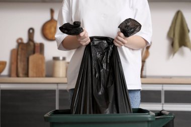 Mutfaktaki çöp kutusundan bir çanta dolusu çöp alan kadın.