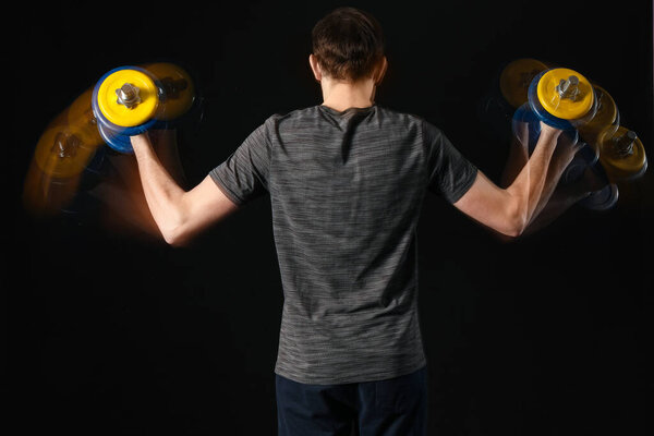 Young male fitness trainer exercising with dumbbells in motion on black background, back view