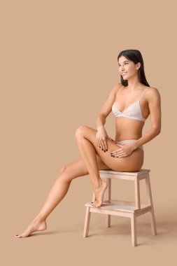 Selülit sorunu olan genç bir kadın bej arka planda taburede oturuyor.