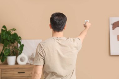 Oturma odasındaki bej duvarın yanında klimalı uzaktan kumandalı yakışıklı bir genç.