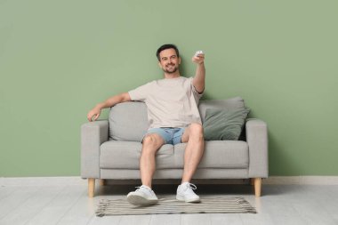 Yakışıklı genç adam yeşil duvarın yanındaki kanepede otururken klimayı açıyor.