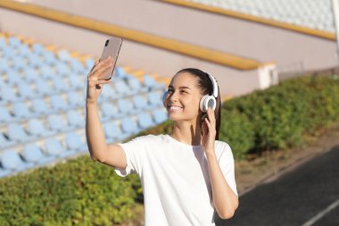 Stadyumda cep telefonu ve kulaklıkları olan genç bir kadın selfie çekiyor.