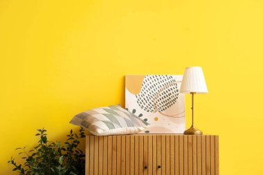 Ahşap dolap, masa lambası, yastık ve sarı duvarın yanında boya.
