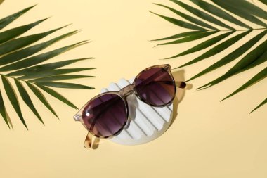 Soluk sarı arka planda şık bir güneş gözlüğü ve palmiye yapraklarıyla dur.