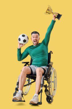Sarı arka planda top ve altın bardakla tekerlekli sandalyede oturan erkek futbolcu.