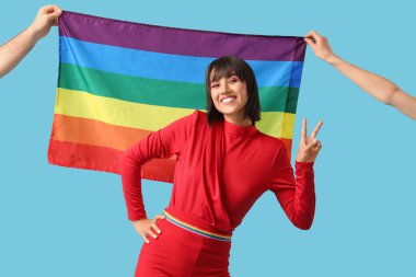 Güzel bir genç kadın mavi arka planda LGBT bayrağı ile zafer jesti ve eller gösteriyor.