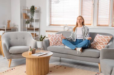 Genç ve güzel bir kadın oturma odasında rahat bir kanepede meditasyon yapıyor.