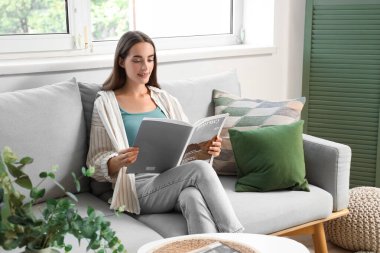 Oturma odasındaki gri koltukta dergi okuyan genç bir kadın.