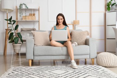 Genç kadın oturma odasındaki gri koltukta dizüstü bilgisayar kullanıyor.