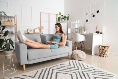 Genç kadın oturma odasındaki gri koltukta dizüstü bilgisayar kullanıyor.