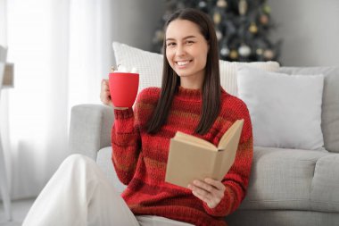 Evdeki Noel ağacının yanında elinde kakao bardağıyla kitap okuyan mutlu genç bir kadın.