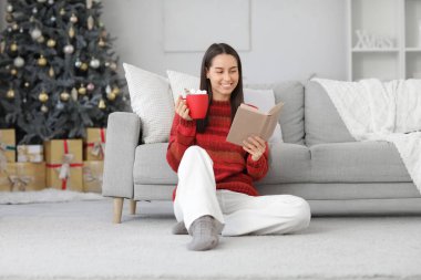 Evdeki Noel ağacının yanında elinde kakao bardağıyla kitap okuyan mutlu genç bir kadın.