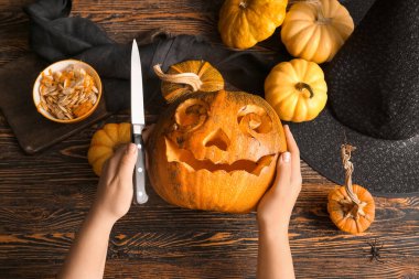 Kahverengi ahşap arka planda Cadılar Bayramı için balkabağı oyan cadı şapkalı ve bıçaklı kadın.