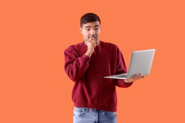 Turuncu arka planda dizüstü bilgisayarı olan korkmuş genç bir adam.