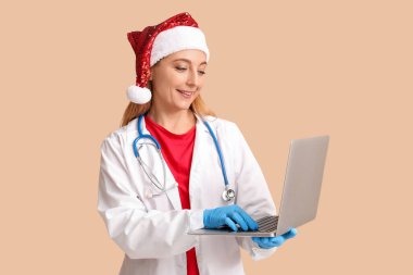 Bej arka planda modern dizüstü bilgisayarı olan Noel Baba şapkalı olgun bir kadın doktor.