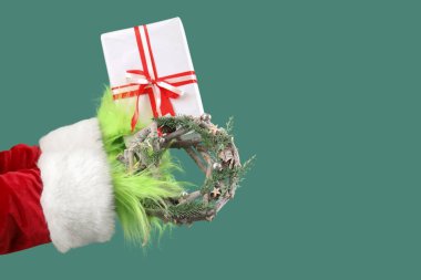 Hairy hands of creature in Santa costume with Christmas gift box and wreath on green background clipart