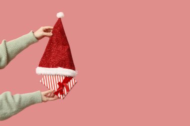 Pembe arka planda Noel Baba şapkası ve hediye kutusu tutan kadın eller.