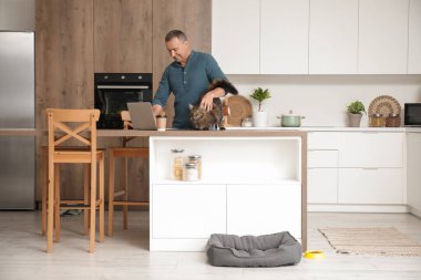 Evde dizüstü bilgisayarı ve kahve fincanı olan olgun bir adam masadaki sevimli kediyi okşuyor.